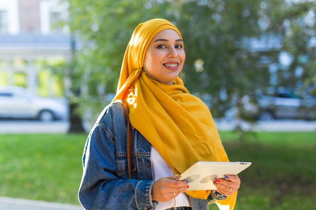 Estudante árabe. Linda aluna muçulmana usando o tablet de exploração do hijab amarelo brilhante.