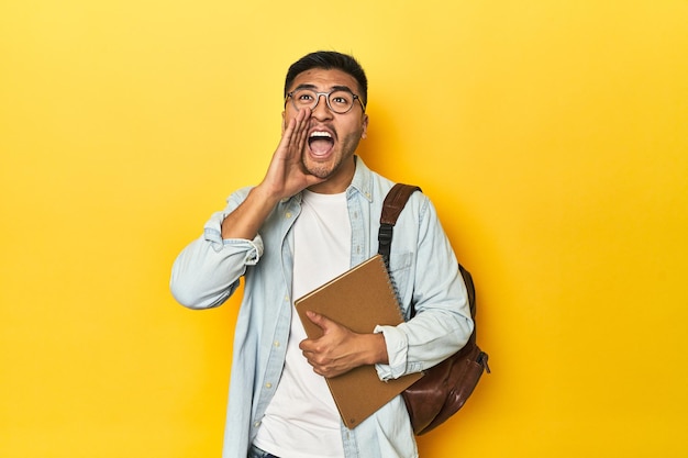 Estudante asiático com mochila e caderno com fundo de estúdio amarelo gritando