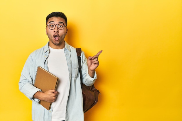 Estudante asiático com mochila e caderno fundo de estúdio amarelo apontando para o lado