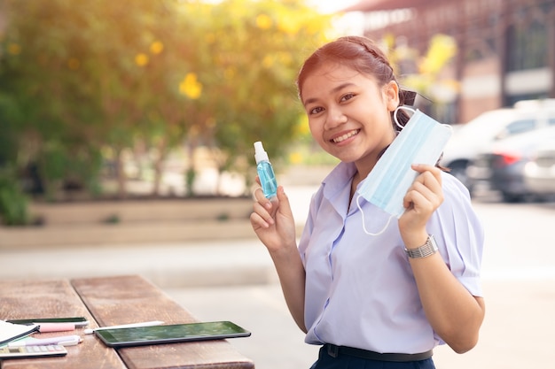 Estudante asiático saudável feliz usando máscara facial e spray de álcool na escola para proteção do vírus corona (covid-19).