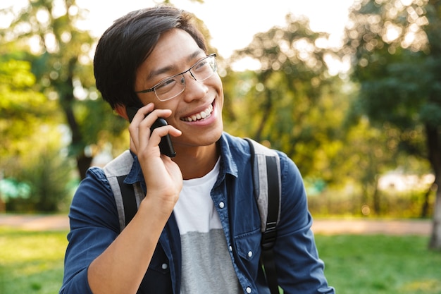 Estudante asiático sorridente em óculos falando por um smartphone e desviando o olhar enquanto está no parque