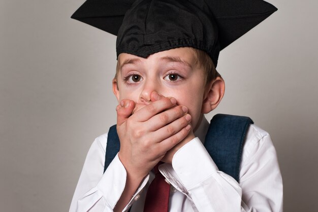 Estudante assustado cobriu a boca com as mãos. Menino com chapéu de estudante. Fundo branco. Ensino fundamental.