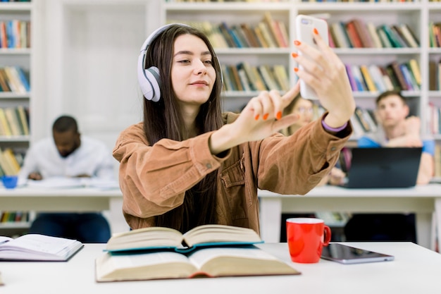 Estudante atraente jovem feliz, vestindo roupas casuais e fones de ouvido hipster, sentado a mesa com livros, segurando o telefone móvel para foto de selfie