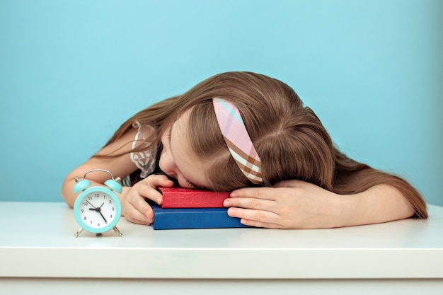 Estudante cansada dorme sobre seus livros ao lado de um relógio sobre fundo azul claro.