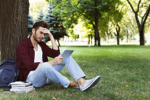 Estudante concentrado se preparando para o exame no parque. Homem casual com tablet digital, sentado na grama, copie o espaço. Tecnologia, comunicação, educação e conceito de trabalho remoto.