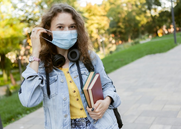 Estudante de cabelo encaracolado com máscara médica facial segurando uma pilha de livros ao ar livre Educação durante uma pandemia
