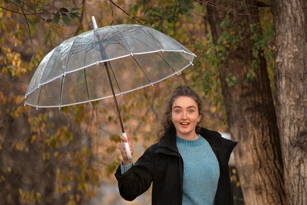 Estudante de casaco preto e com guarda-chuva transparente posa para foto Garota de cabelo escuro caminha no parque outono