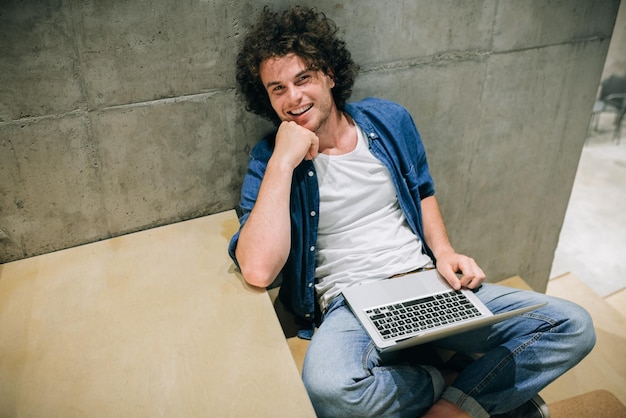 Foto estudante de homem alegre com cabelos cacheados usando laptop para aprender navegação on-line na internet sentado nas escadas na parede de concreto sorrindo masculino lendo e digitando em seu laptop no escritório