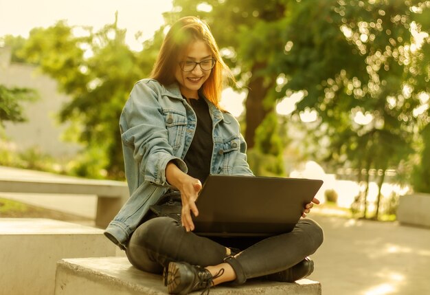 Estudante de jovem mulher feliz em uma jaqueta jeans, sentado no parque e olha para a tela do laptop. Ensino à distância. Chamada online. Conceito moderno da juventude.