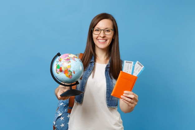 Estudante de mulher jovem bonita de óculos com a mochila segurando a luva mundial, passaporte, bilhetes de cartão de embarque isolados sobre fundo azul. Educação em faculdade universitária no exterior. Conceito de voo de viagens aéreas.