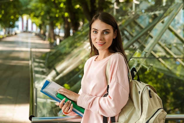 Foto estudante de mulher jovem e atraente em pé ao ar livre no parque da cidade, segurando livros, carregando uma mochila