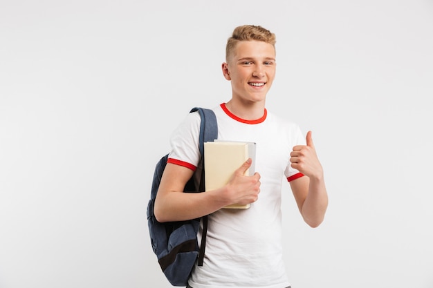 estudante do sexo masculino feliz usando mochila, sorrindo e aparecendo o polegar enquanto segura livros isolados no branco