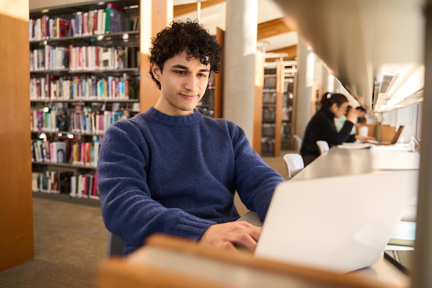 Estudante do sexo masculino latino-americano usando laptop fazendo pesquisas na internet na biblioteca de inovação moderna
