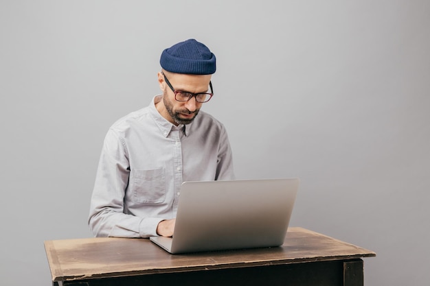 Estudante do sexo masculino na moda publica no computador laptop se prepara para o exame passa tempo na educação e obtém conhecimento posa na mesa antiga usa óculos de chapéu elegantes e camisa branca