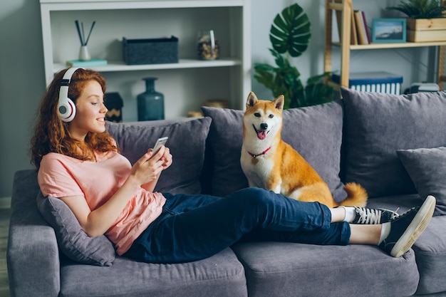 Estudante em fones de ouvido ouvindo música usando smartphone no sofá com cachorro