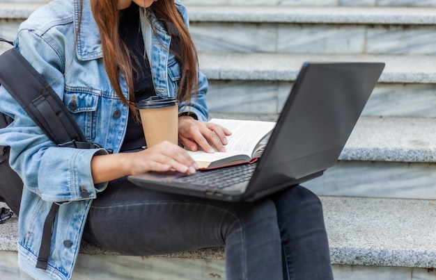 Estudante feliz mulher moderna na jaqueta jeans e mochila sentado na escada e usa laptop com livro ao ar livre. Ensino à distância. Conceito de juventude moderna.