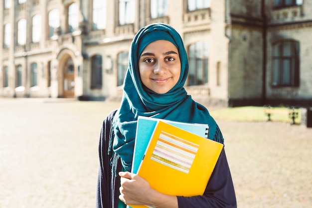 Estudante fêmea novo muçulmano de sorriso que guarda os livros que estão a faculdade próxima. Feliz garota árabe em hijab. Mulher asiática no treinamento