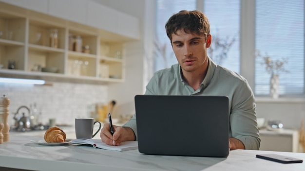 Estudante focado estudando on-line sentado na cozinha em close-up homem trabalhando em laptop