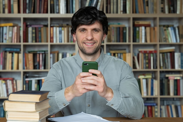 Estudante homem usando smartphone, aprendendo sentado lendo livros na biblioteca da universidade