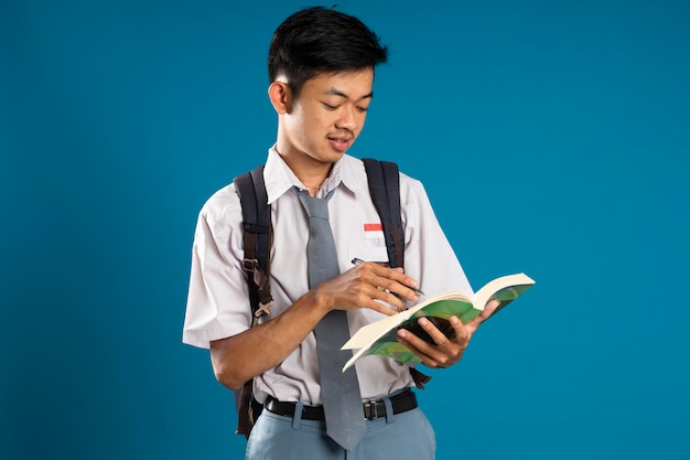 Estudante indonésio do ensino médio pensando enquanto olha para o livro que ele está carregando sobre fundo azul