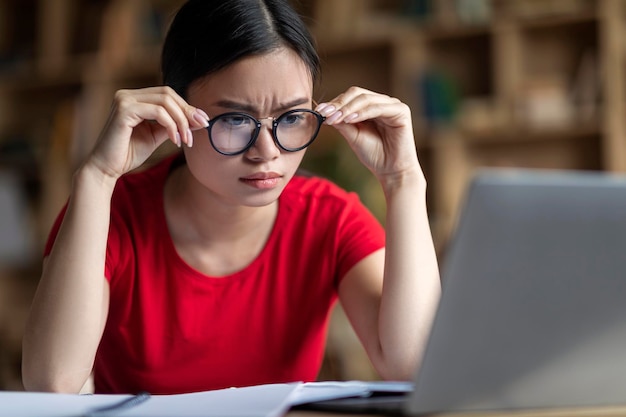 Estudante japonesa séria e inteligente em óculos assistindo aula on-line no laptop no interior da sala