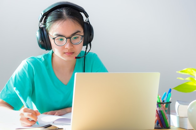 Estudante jovem asiática com fones de ouvido de óculos estuda uma nota escrita séria em um livro procurando uma videoconferência em um computador laptop aula universitária online Internet aprendizagem educação a distância em casa