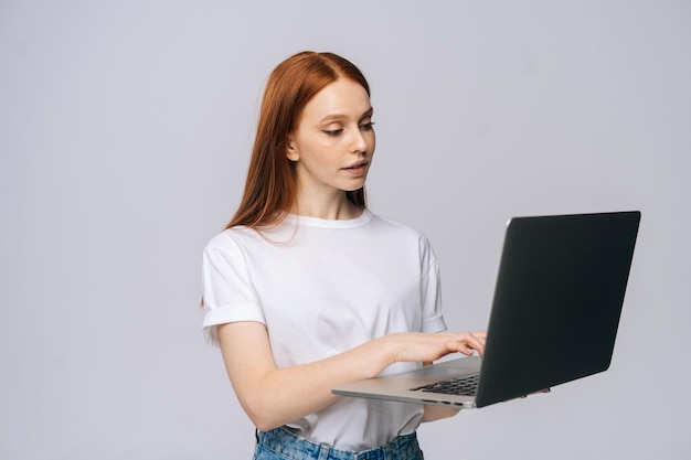 Estudante jovem bem sucedida segurando o computador portátil e digitando no fundo cinza isolado. Modelo de senhora bonita ruiva mostrando emocionalmente expressões faciais no estúdio, copie o espaço.