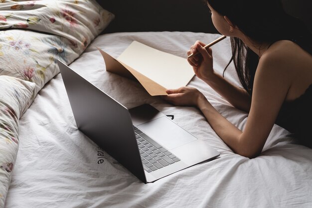 Estudante jovem deita-se na cama com laptop e notebook e faz anotações. educação a distância