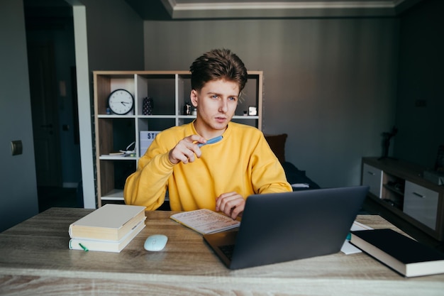 Estudante jovem inteligente estuda remotamente em casa se senta em uma mesa com um laptop e livros escreve com uma caneta em um caderno e desvia o olhar com um rosto pensativo Ensino à distância na escola