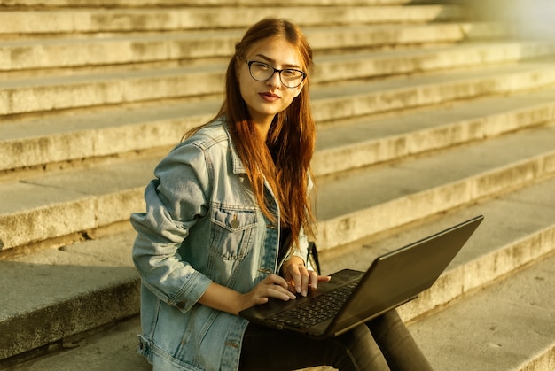 Estudante jovem moderna com uma jaqueta jeans, sentada na escada com o laptop. Ensino à distância. Conceito moderno da juventude.