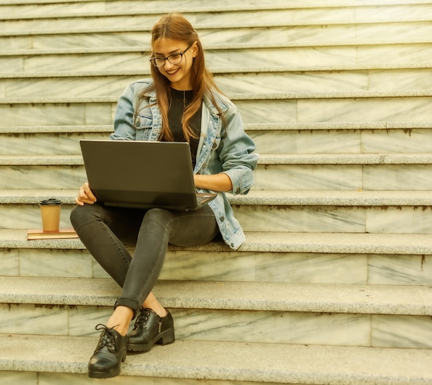 Estudante jovem moderna com uma jaqueta jeans, sentada na escada e assistindo a um vídeo no laptop. Ensino à distância. Conceito moderno da juventude.