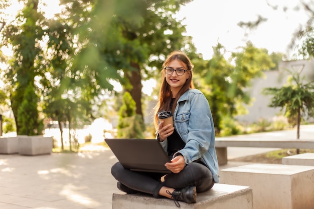 Estudante jovem moderno em uma jaqueta jeans, sentado no parque e usa o laptop com uma xícara de café na mão. ensino à distância. conceito de juventude moderna.