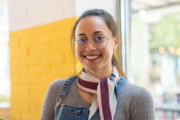Estudante jovem nerd latina sorrindo para a câmera dentro de casa na sala de aula universidade Garota com óculos contra o fundo da parede amarela Foco no rosto