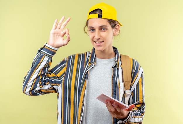 Estudante jovem sorridente usando mochila com tampa segurando um caderno mostrando um gesto de ok