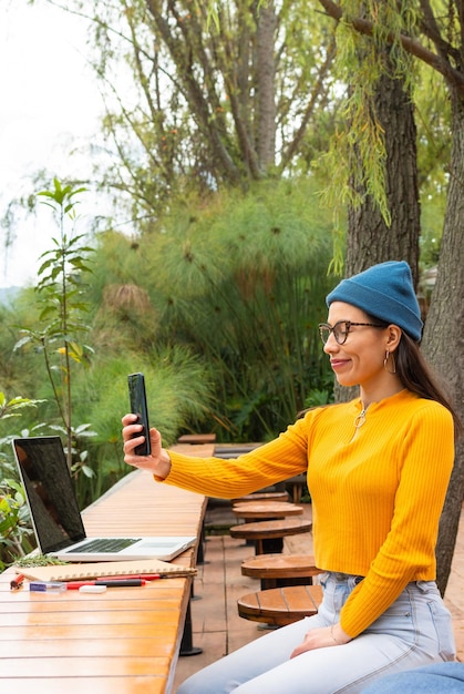 Estudante latina sentada em um parque público fazendo um vídeo para redes sociais com seu celular na frente de seu laptop e seu material de estudo