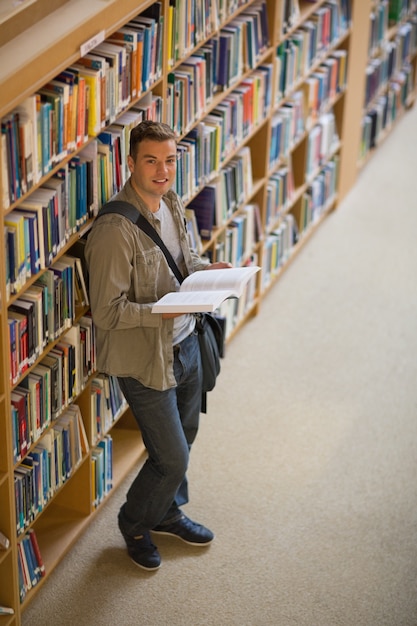 Estudante lendo um livro em pé na biblioteca sorrindo para a câmera