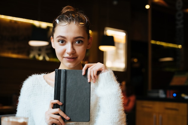 Estudante linda garota segurando um livro, sentada em um café e sorrindo