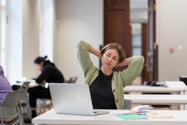 Estudante madura cansada fazendo uma pausa esticando os braços enquanto estuda on-line na biblioteca da universidade