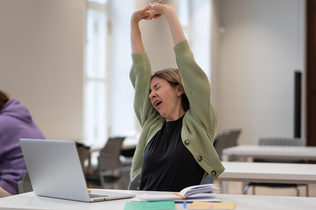 Foto estudante madura cansada fazendo uma pausa esticando os braços enquanto estuda on-line na biblioteca da universidade