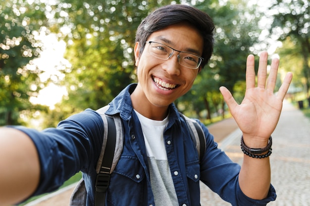 Estudante masculino asiático sorridente em óculos fazendo selfie e acenando para a câmera ao ar livre