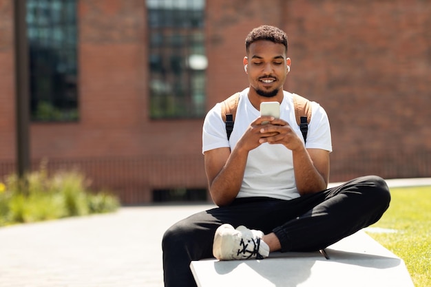 Estudante negro na moda usando mensagens de texto para smartphone e ouvindo música em fones de ouvido sentados ao ar livre na área urbana