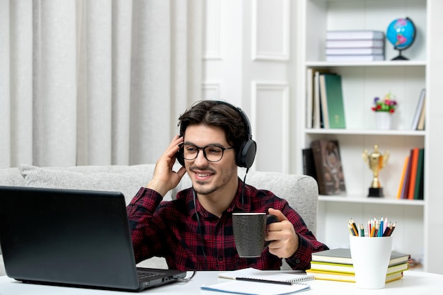 Estudante on-line bonitinho de camisa xadrez com óculos estudando no computador sorrindo e ouvindo