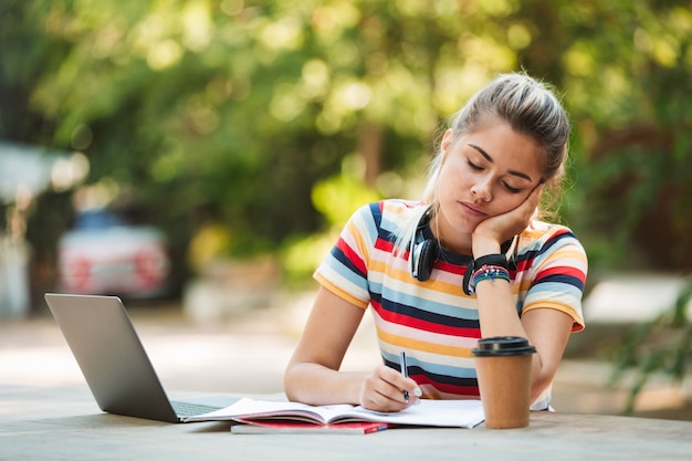 Estudante pensativo jovem bonito sentado no parque usando o computador portátil escrevendo notas.