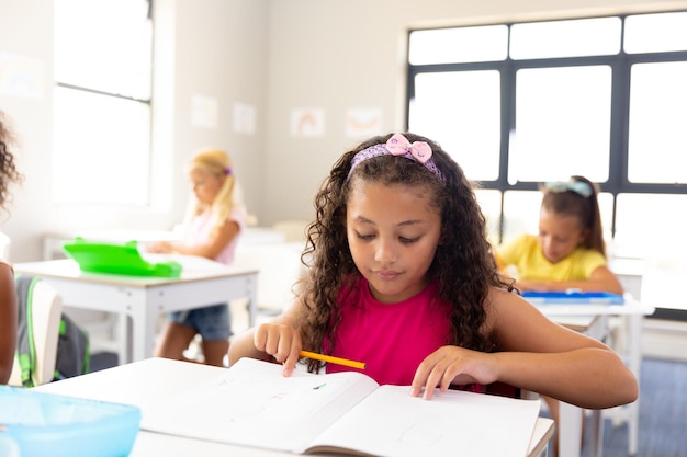 Foto estudante primária biracial lendo livro enquanto está sentada na mesa em sala de aula