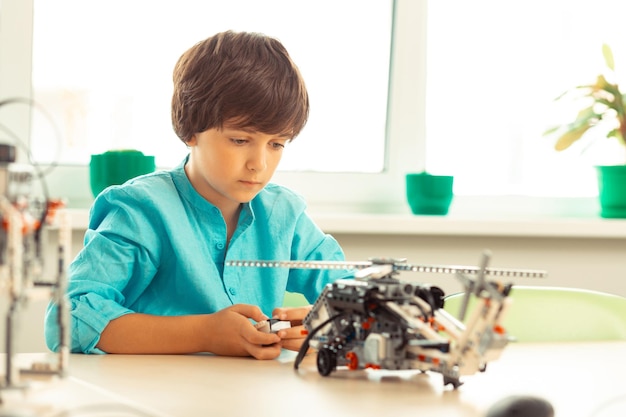 Estudante primário concentrado sentado na sala de aula com controle remoto nas mãos ligando um modelo de helicóptero