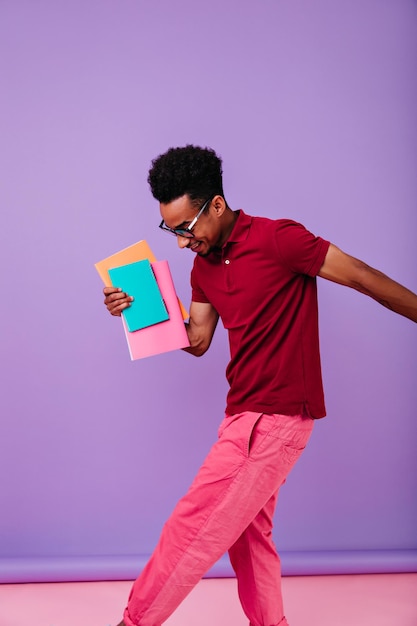 Estudante satisfeito em óculos dançando após os exames Foto interna de um jovem africano feliz com livros posando em fundo roxo