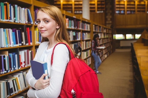 Estudante sorridente com mochila segurando um livro na biblioteca