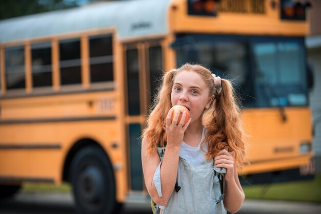 Estudante sorridente da educação pronta para embarcar no ônibus escolar na frente do ônibus escolar comendo maçã