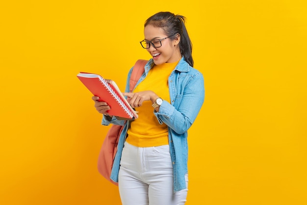 Estudante sorridente em roupas jeans com mochila lendo no caderno de trabalho isolado em fundo amarelo Educação no conceito de faculdade universitária do ensino médio