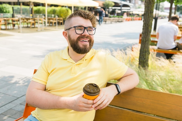 Foto estudante sorridente milenar em camisa amarela, óculos, sentado no banco, a beber café.